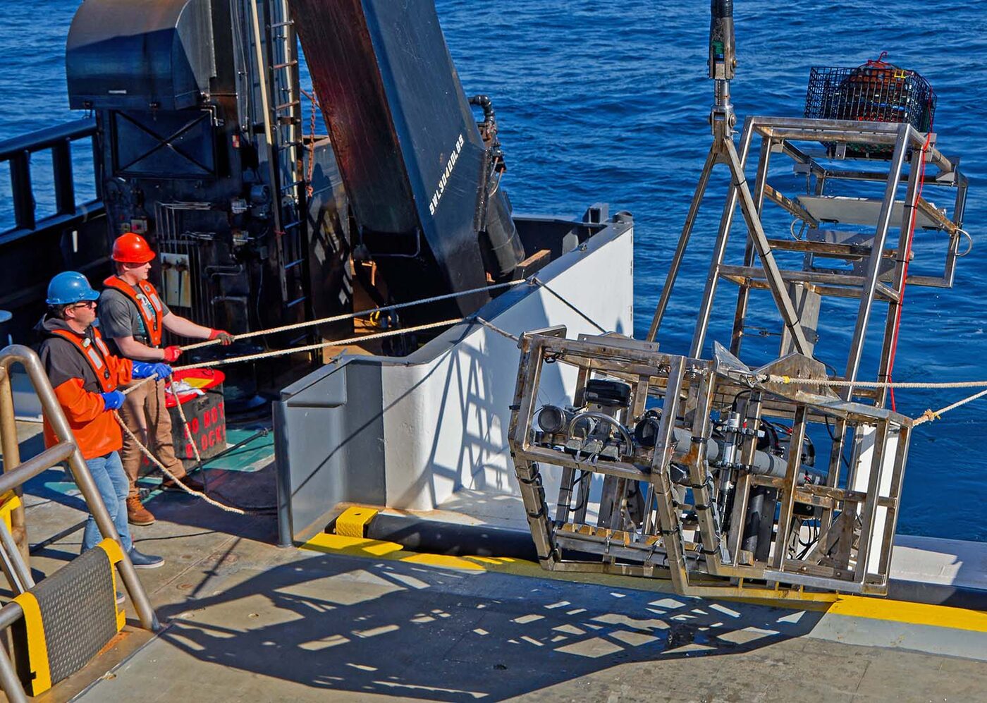 Two crew members guiding the Habitat Camera frame over the stern of a NOAA research vessel